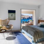Dormitorio con cabecero de madera, ropa de cama azul, butaca de ratán y salida a la terraza