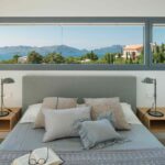 Bedroom with wide horizontal window above the bed, sea and mountain views, grey cushions and a book