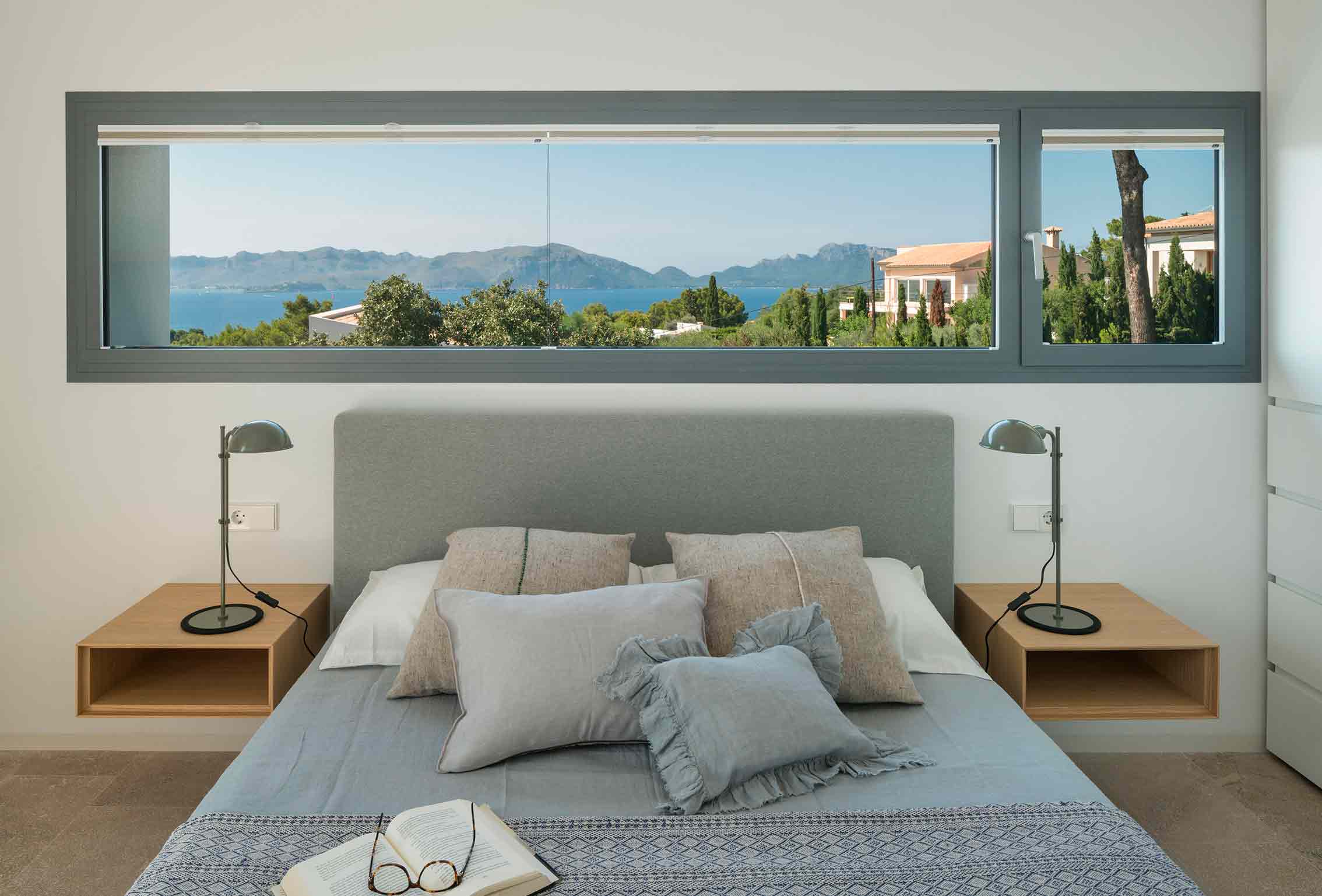 Bedroom with wide horizontal window above the bed, sea and mountain views, grey cushions and a book
