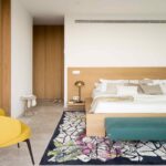 Dormitorio con cama doble, cabecero de roble, butaca amarilla, alfombra estampada y banco turquesa