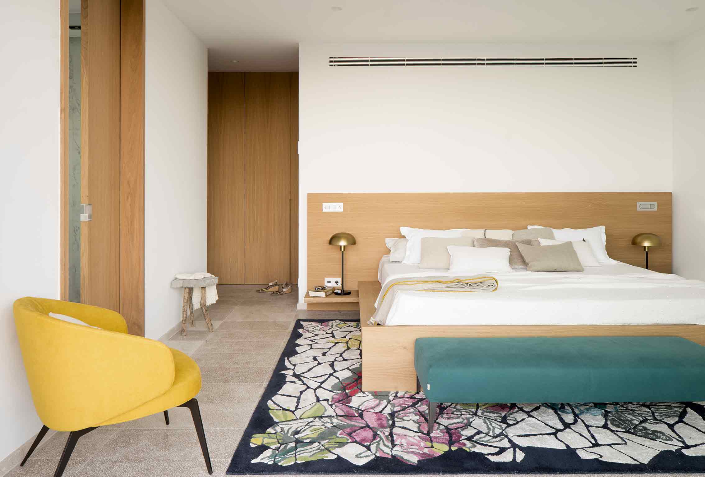 Dormitorio con cama doble, cabecero de roble, butaca amarilla, alfombra estampada y banco turquesa