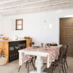 Esszimmer mit weißem Tisch, gemusterter Tischdecke, Eisenstühlen, weiß getünchten Balken und Holzküche