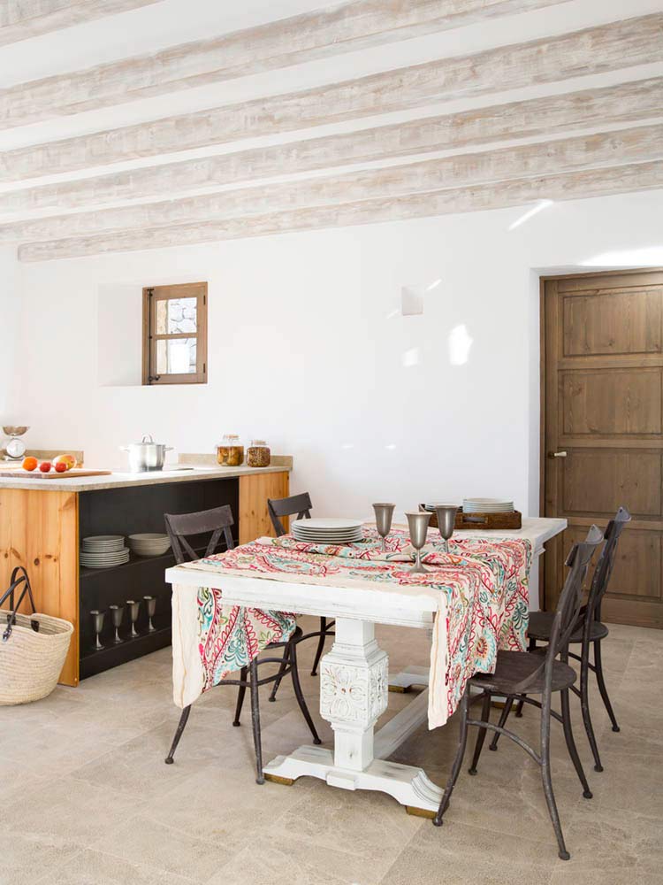 Esszimmer mit weißem Tisch, gemusterter Tischdecke, Eisenstühlen, weiß getünchten Balken und Holzküche