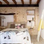 Schlafzimmer mit freiliegender Steinwand, Holzbalken, geblümter Tagesdecke und Einbauregalen