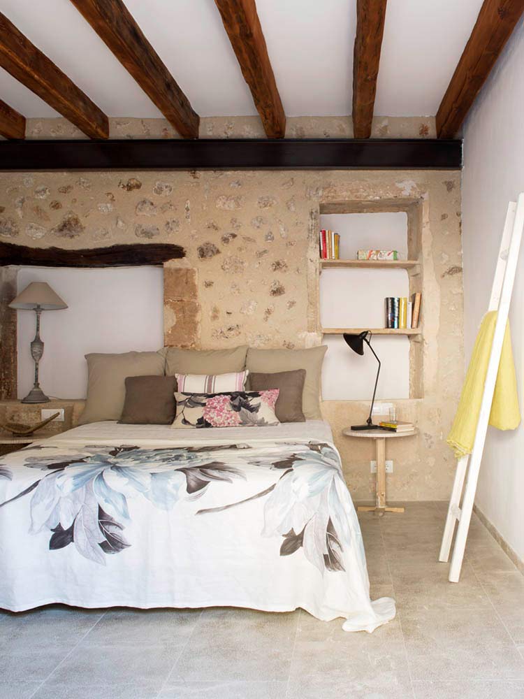 Schlafzimmer mit freiliegender Steinwand, Holzbalken, geblümter Tagesdecke und Einbauregalen