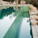 Pool mit grünem Mosaik und Spiegelungen, Felsen rechts und Rattanstühlen im Hintergrund