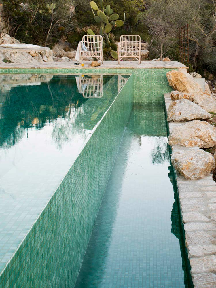 Pool mit grünem Mosaik und Spiegelungen, Felsen rechts und Rattanstühlen im Hintergrund