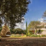 Jardín con cama colgante bajo una encina y la casa al fondo tras un muro de piedra