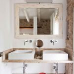 Baño con dos lavabos blancos sobre encimera de piedra, grifos de pared y espejo de marco envejecido