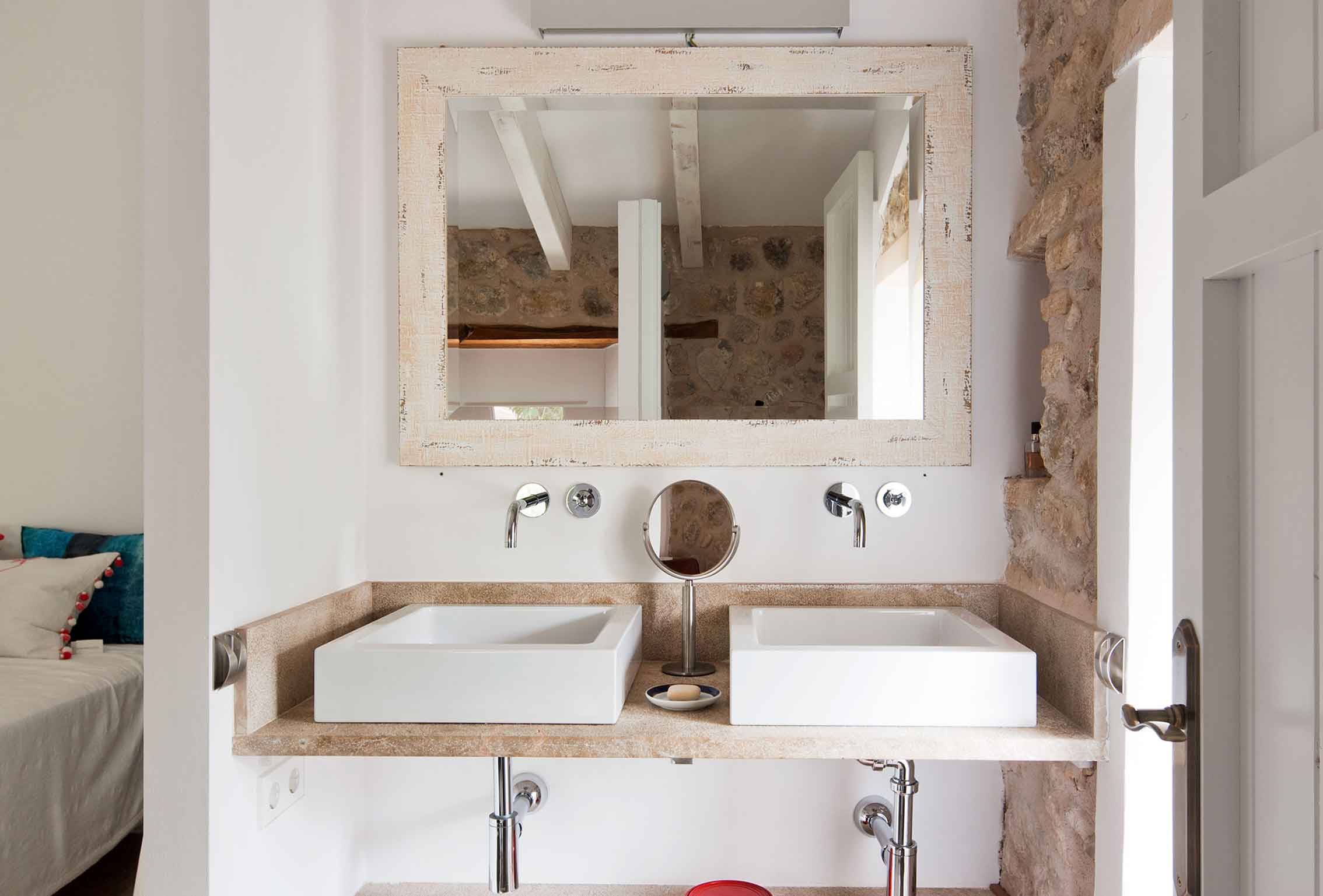 Baño con dos lavabos blancos sobre encimera de piedra, grifos de pared y espejo de marco envejecido