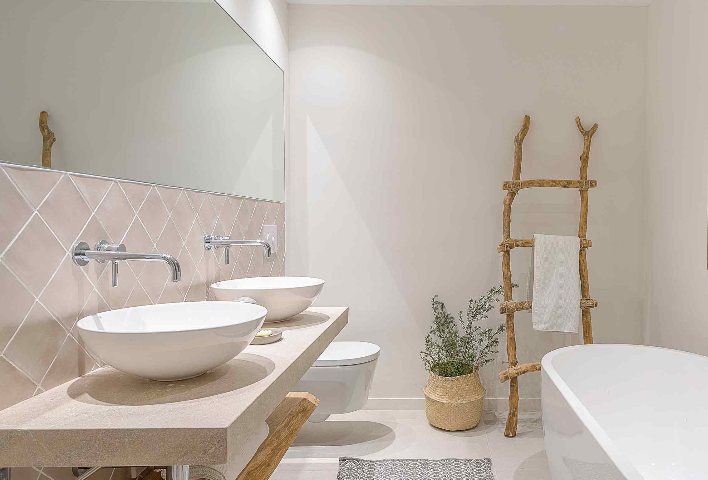 Bad mit diagonal verlegten rosa Fliesen, zwei Waschbecken, freistehender Badewanne und rustikaler Handtuchleiter aus Ästen