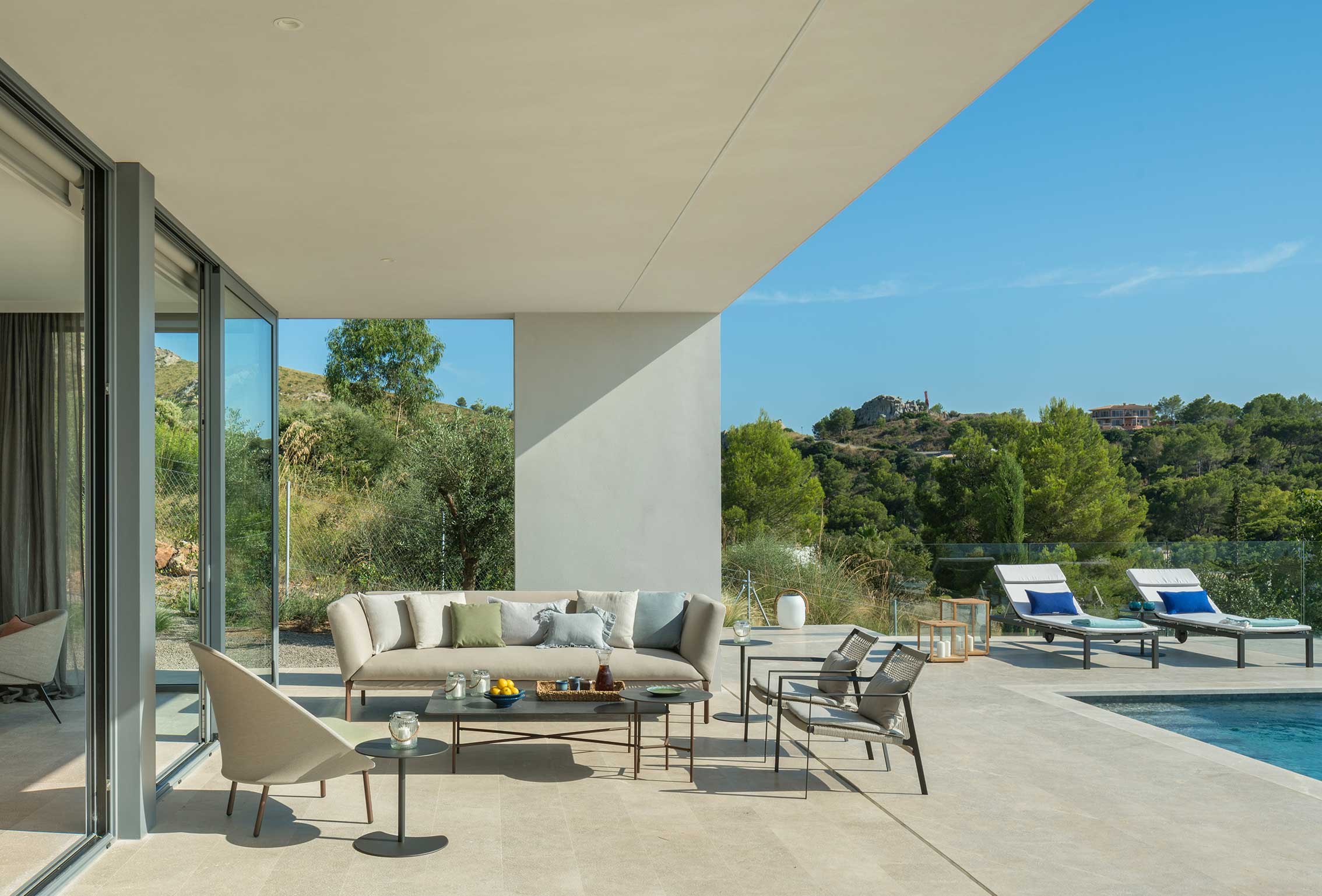 Light stone terrace with sofa, armchairs and sun loungers beside the pool, with hillside beyond