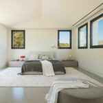 Weißes Schlafzimmer mit niedrigem Bett, hellem Teppich, grauer Ottomane und dunkel gerahmten Fenstern zum Wald