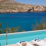 Piscina turquesa con tumbonas blancas, toallas de franja azul y sombrero de paja frente a la bahía