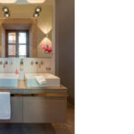 Baño con dos lavabos sobre mueble suspendido gris, espejo con focos y pared de piedra oscura