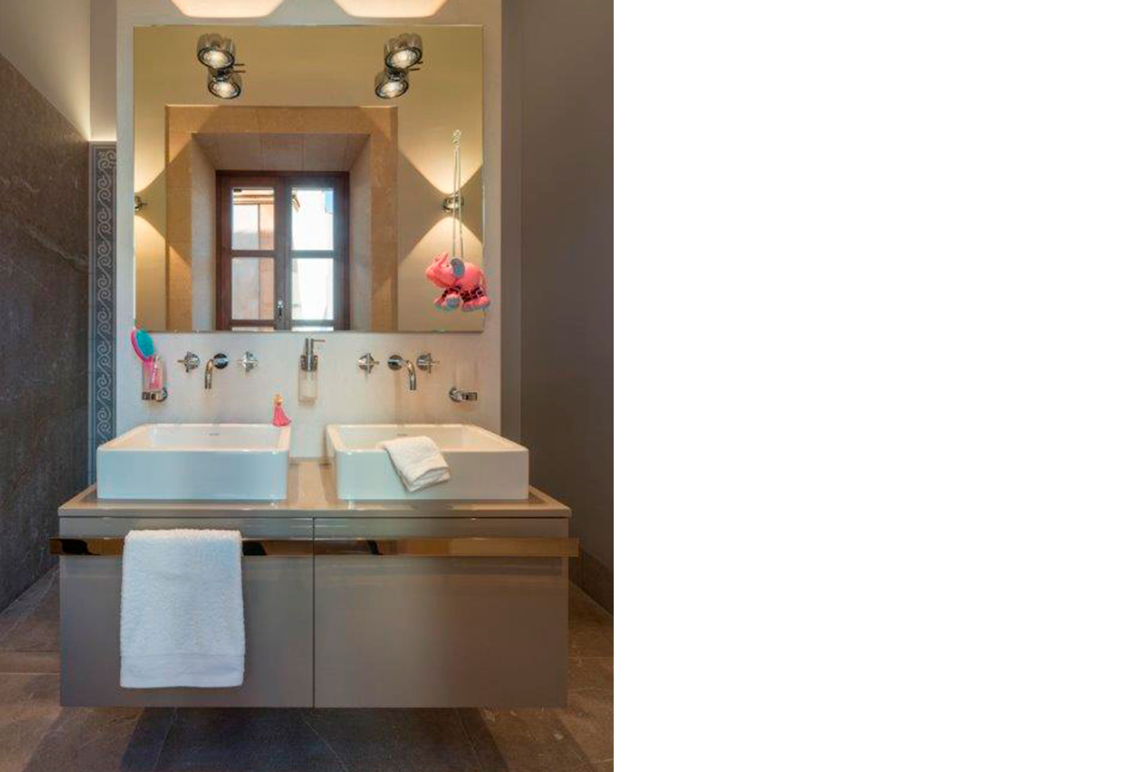Baño con dos lavabos sobre mueble suspendido gris, espejo con focos y pared de piedra oscura