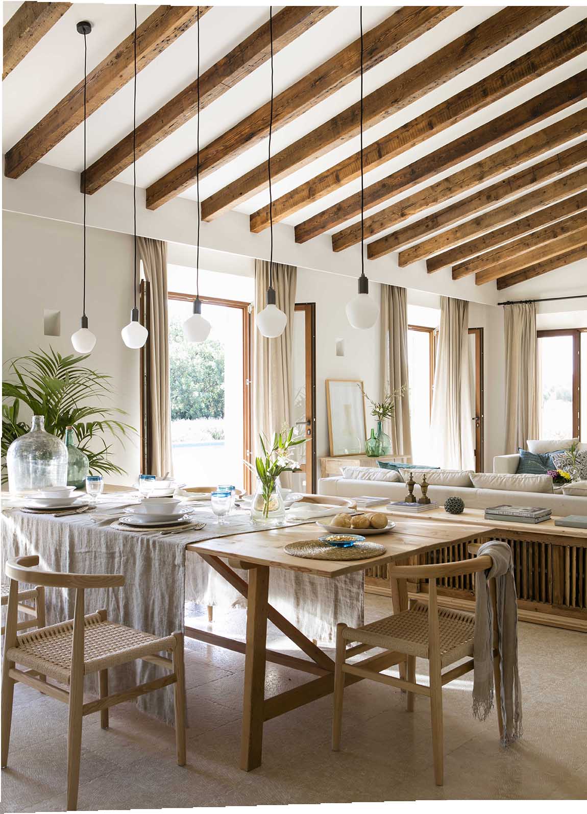 Mesa de comedor con mantel gris, sillas de madera y cuerda, lámparas colgantes y sofá al fondo