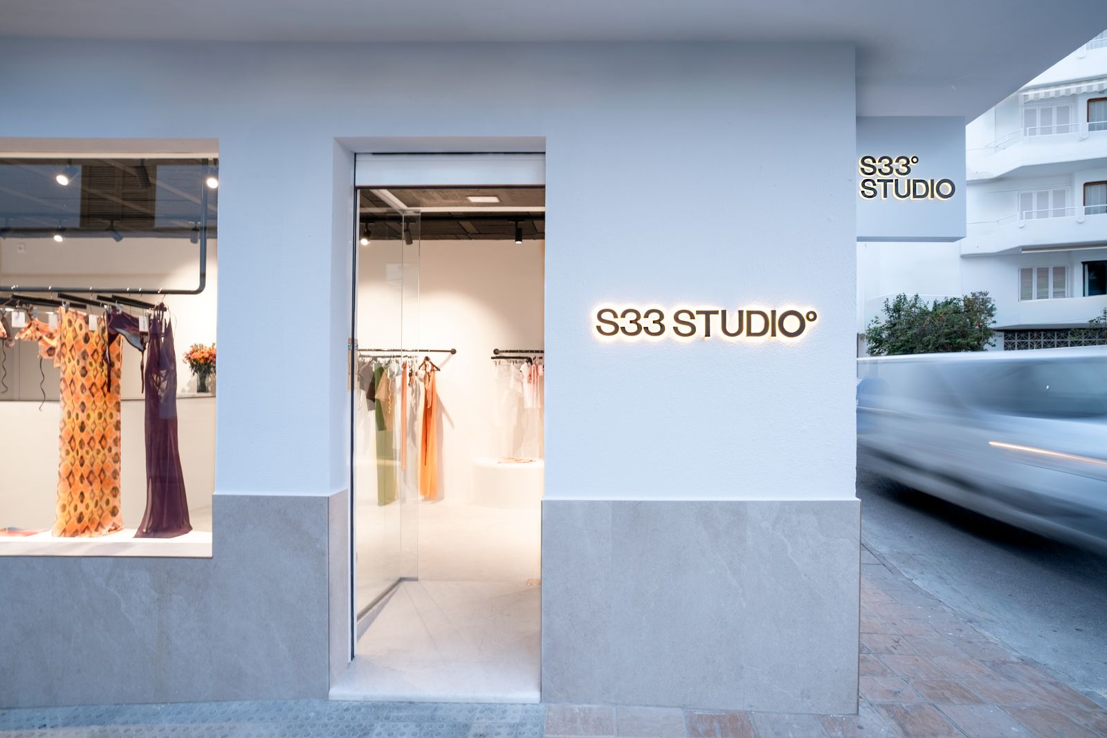 Weiße Fassade von S33 Studio mit beleuchtetem Schriftzug, Schaufenster mit Kleidern und gläserner Tür