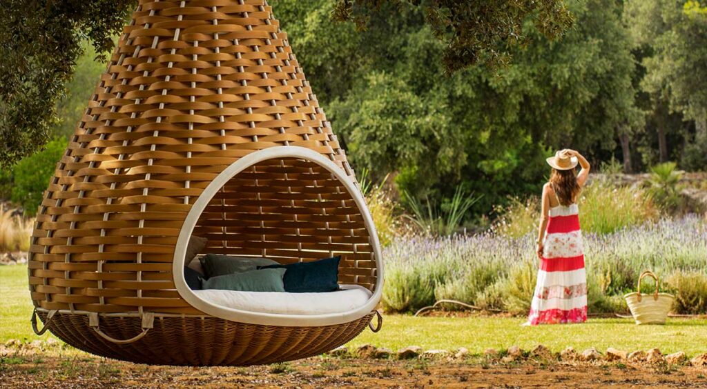 Cama colgante de mimbre trenzado con cojines azules; al fondo lavanda y una mujer con sombrero