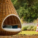 Cama colgante de mimbre trenzado con cojines azules; al fondo lavanda y una mujer con sombrero