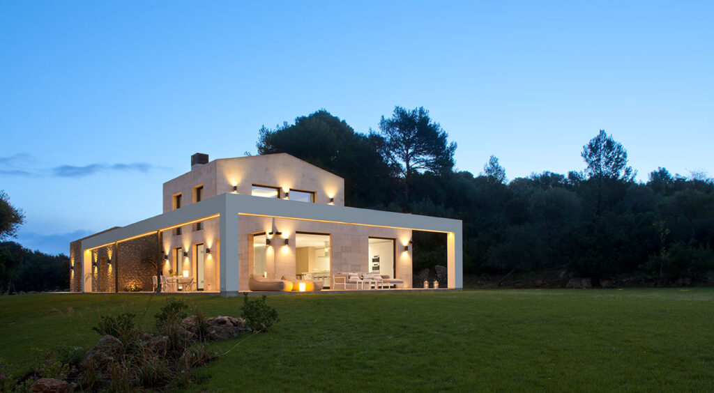 Fachada de la vivienda AC3 al anochecer, con piedra clara, pórtico blanco, luz cálida y jardín de césped
