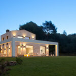 Fachada de la vivienda AC3 al anochecer, con piedra clara, pórtico blanco, luz cálida y jardín de césped