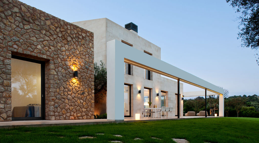 Fachada lateral con muro de piedra irregular, pórtico blanco con luz cálida y terraza con mesa de comedor
