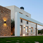 Fachada lateral con muro de piedra irregular, pórtico blanco con luz cálida y terraza con mesa de comedor