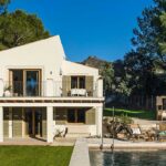 Weiße Fassade der Villa Estrany mit olivgrünen Fensterläden, Balkon, Pool und Kiefern