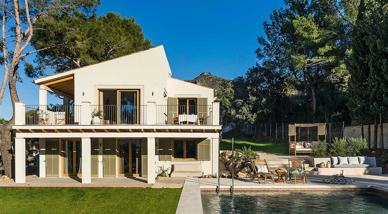Fachada blanca de la villa Estrany con contraventanas verde oliva, balcón, piscina y pinos