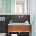 Baño con revestimiento gris oscuro, lavabo sobre mueble de madera, espejo y puertas de cristal translúcido