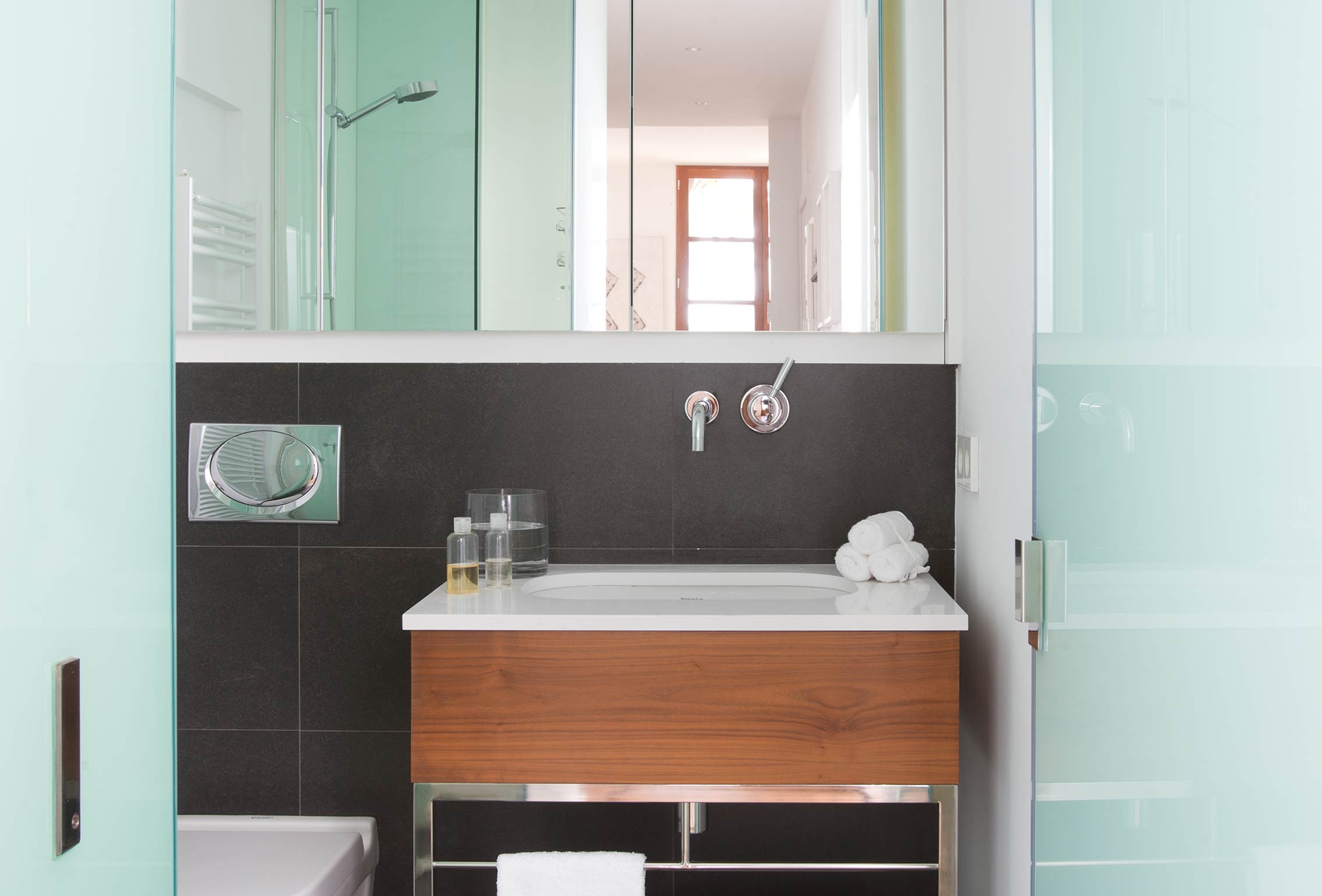Baño con revestimiento gris oscuro, lavabo sobre mueble de madera, espejo y puertas de cristal translúcido