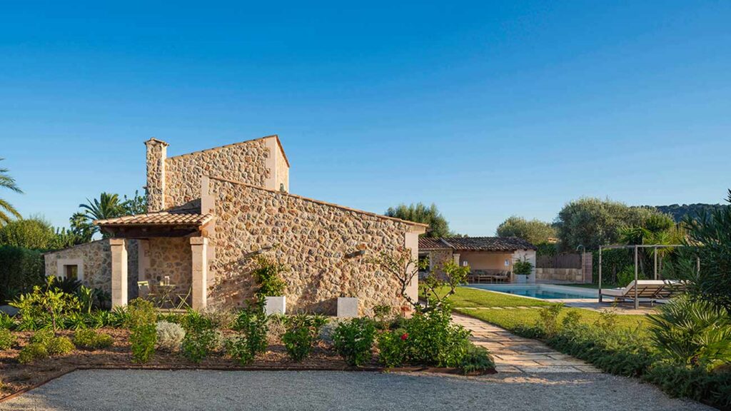 Side of the stone house with a garden of Mediterranean plants and the swimming pool beyond