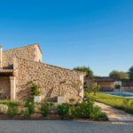 Side of the stone house with a garden of Mediterranean plants and the swimming pool beyond