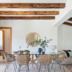 Dining room with a glass table, wicker chairs, round mirror and exposed timber beams