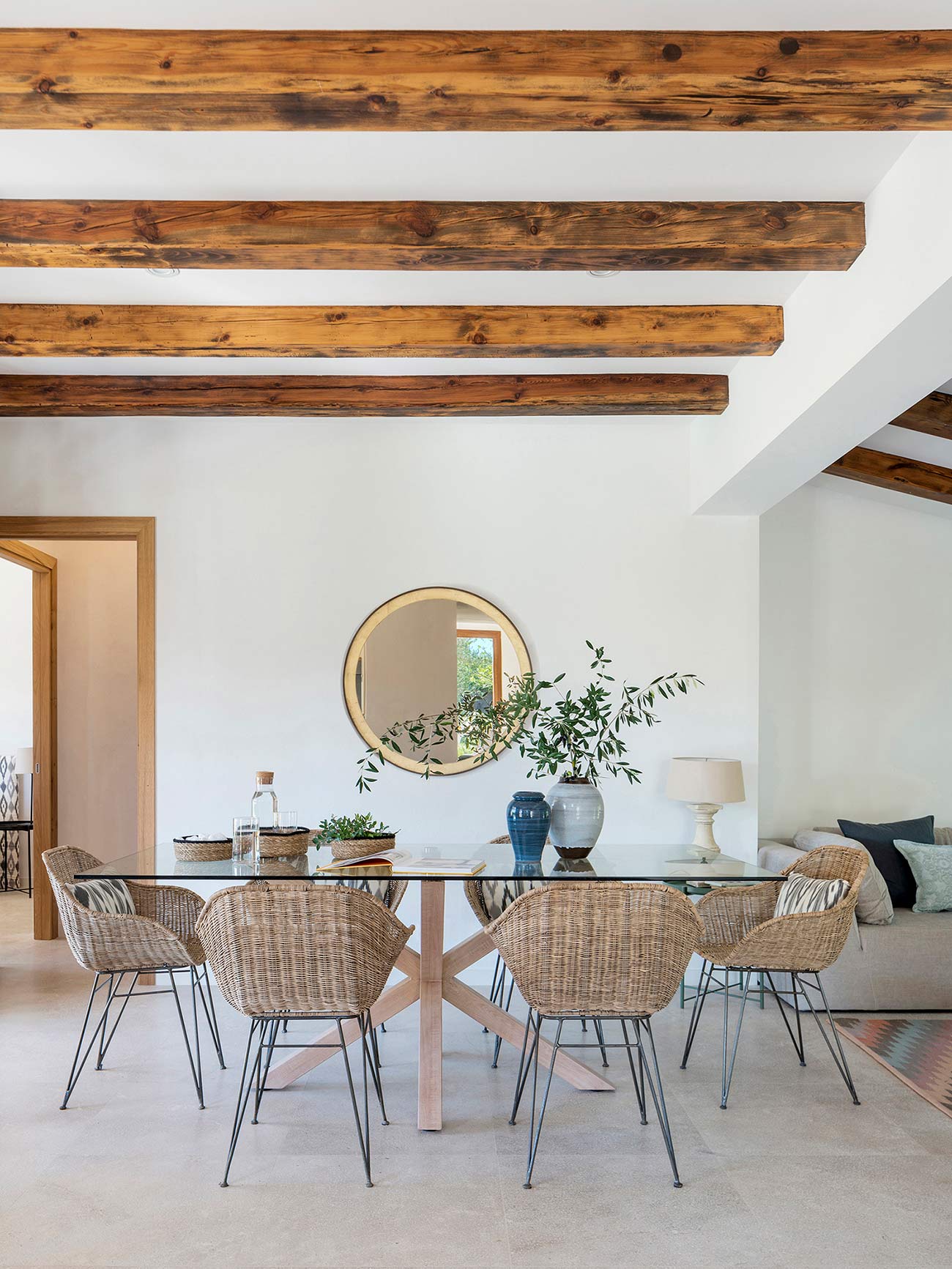 Dining room with a glass table, wicker chairs, round mirror and exposed timber beams