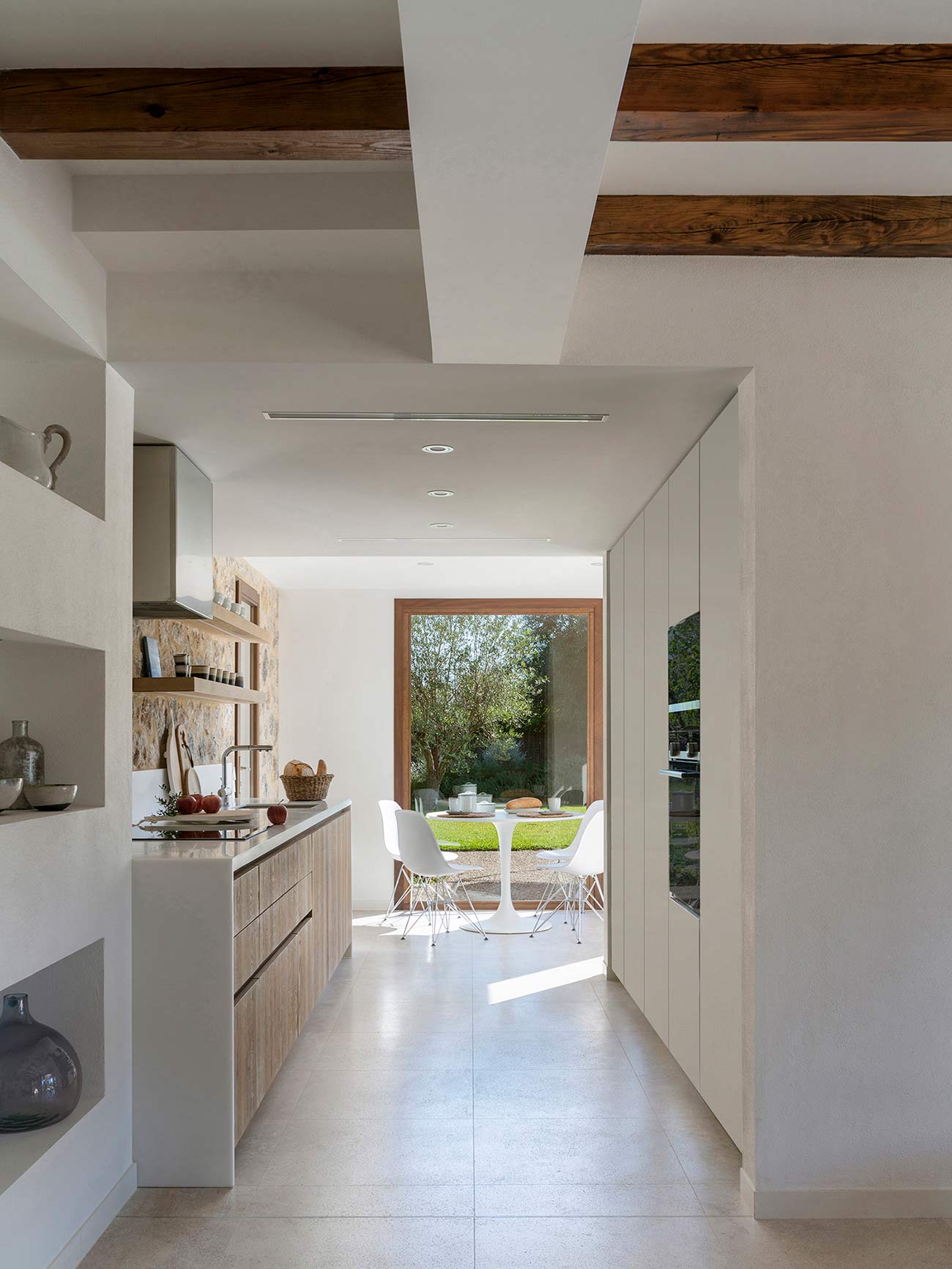 Wood and stone kitchen opening onto a dining room with a white table and a large window facing the garden