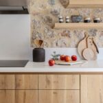 Detalle de cocina con pared de piedra, baldas y armarios de madera clara y encimera blanca