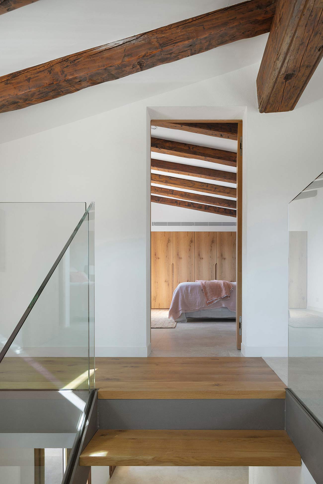 Wooden staircase with a glass balustrade leading to a bedroom with exposed beams
