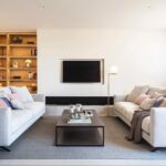 Salón con dos sofás claros enfrentados, estantería de madera iluminada, alfombra gris y televisor empotrado