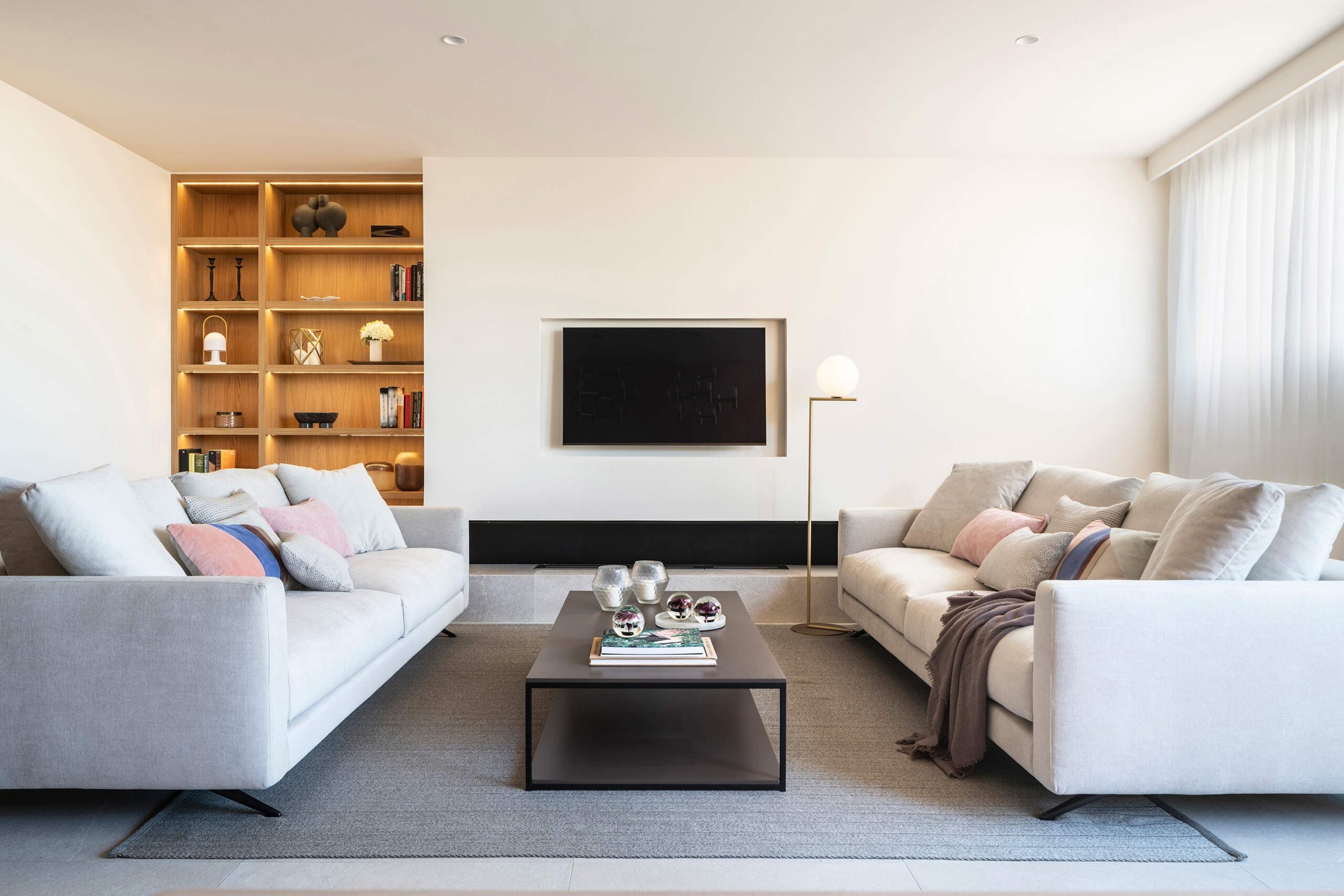 Salón con dos sofás claros enfrentados, estantería de madera iluminada, alfombra gris y televisor empotrado