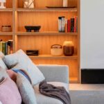 Detalle de estantería de madera iluminada con libros, jarrones y flores blancas junto a un sofá gris