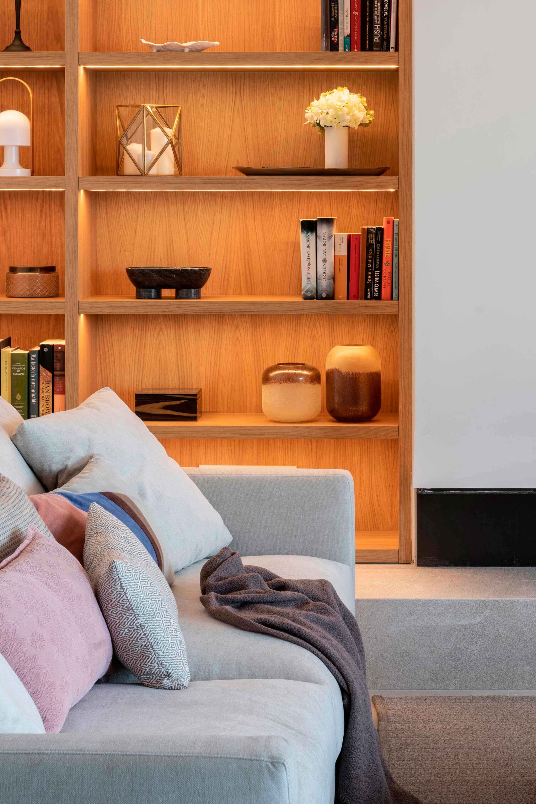 Detalle de estantería de madera iluminada con libros, jarrones y flores blancas junto a un sofá gris