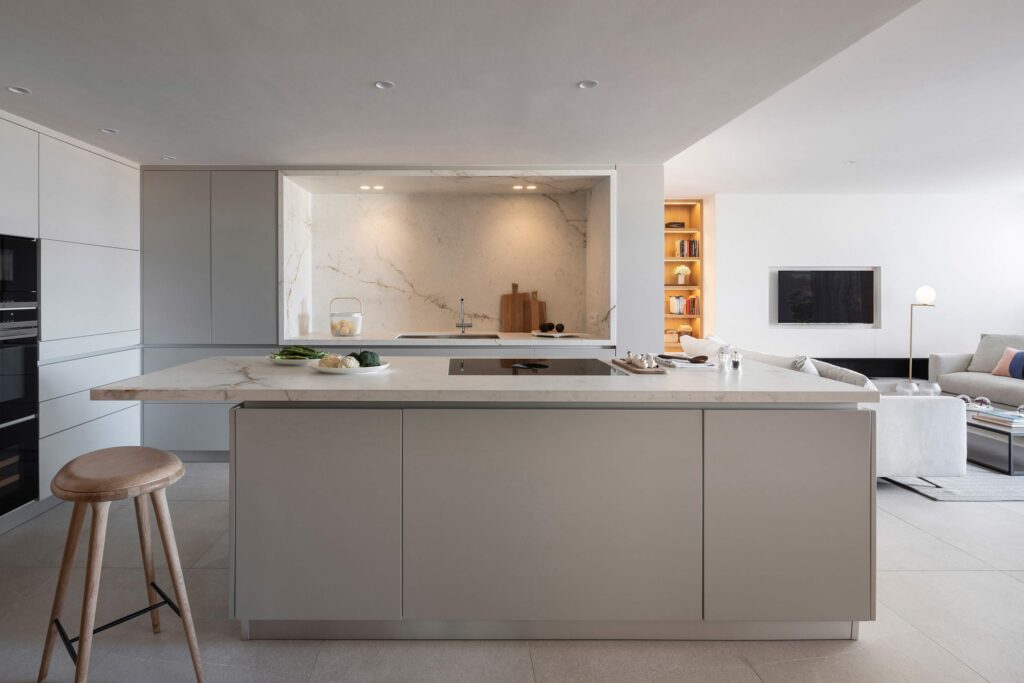 Isla de cocina con encimera clara y taburete de madera, frente marmóreo y salón abierto al fondo