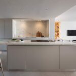 Isla de cocina con encimera clara y taburete de madera, frente marmóreo y salón abierto al fondo