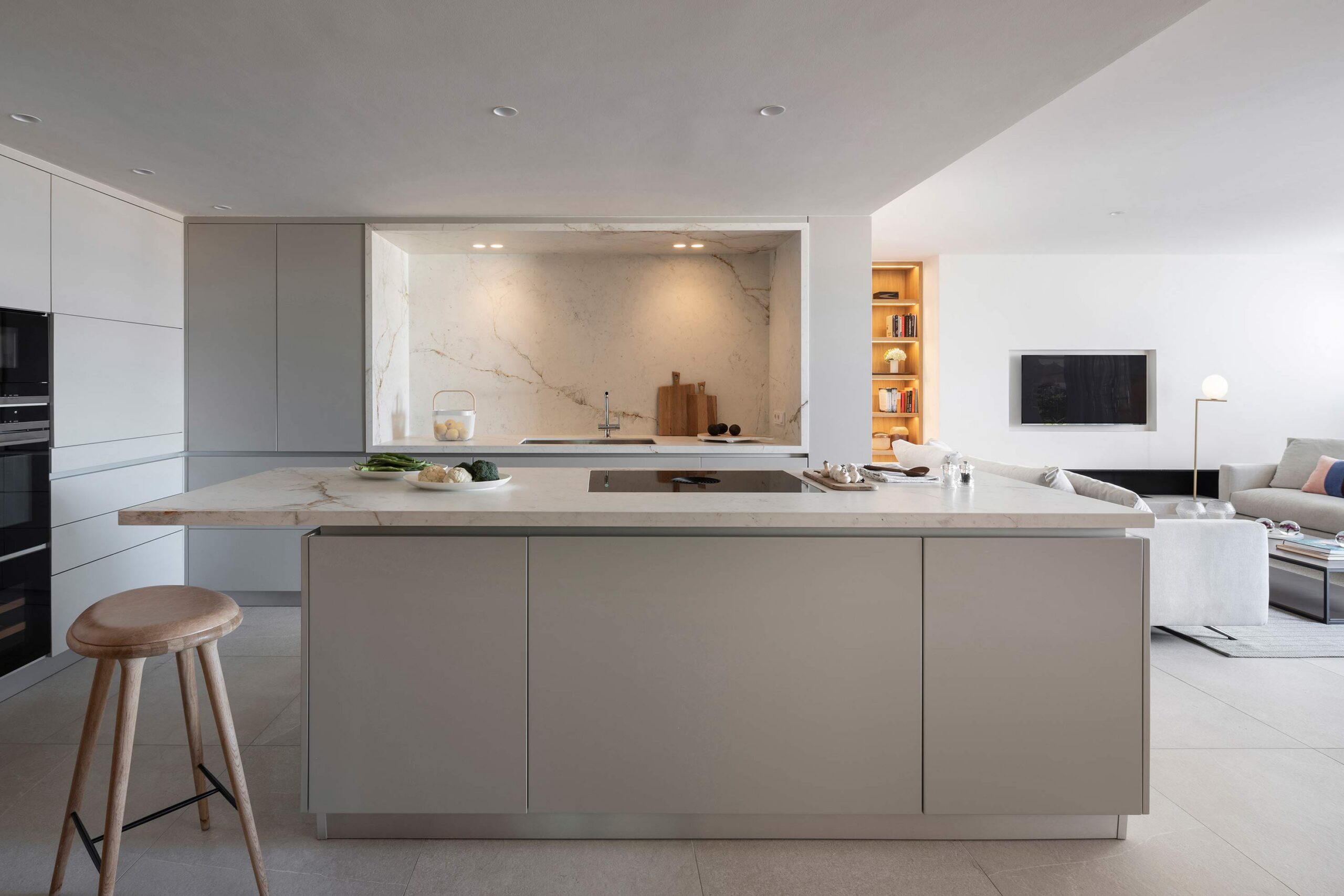 Isla de cocina con encimera clara y taburete de madera, frente marmóreo y salón abierto al fondo