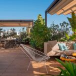 Vista amplia de la terraza con sofá exterior beige, cojines turquesa, butaca blanca, pérgolas y plantas