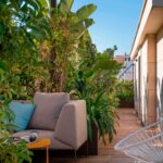 Pasillo de terraza entre plantas tropicales, sofá exterior beige, mesa auxiliar amarilla y butaca blanca bajo cielo azul