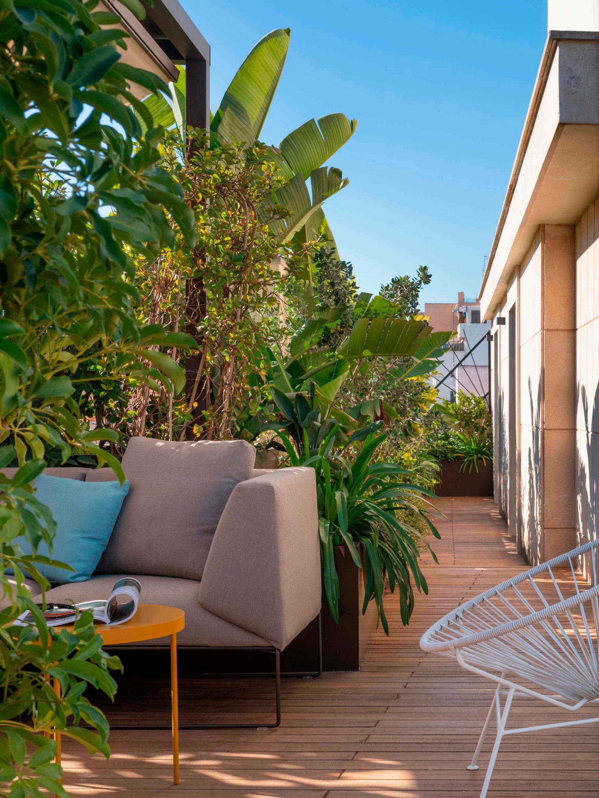 Pasillo de terraza entre plantas tropicales, sofá exterior beige, mesa auxiliar amarilla y butaca blanca bajo cielo azul