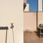 Terraza con ducha exterior en pared beige, silla blanca, barra alta y montañas al fondo